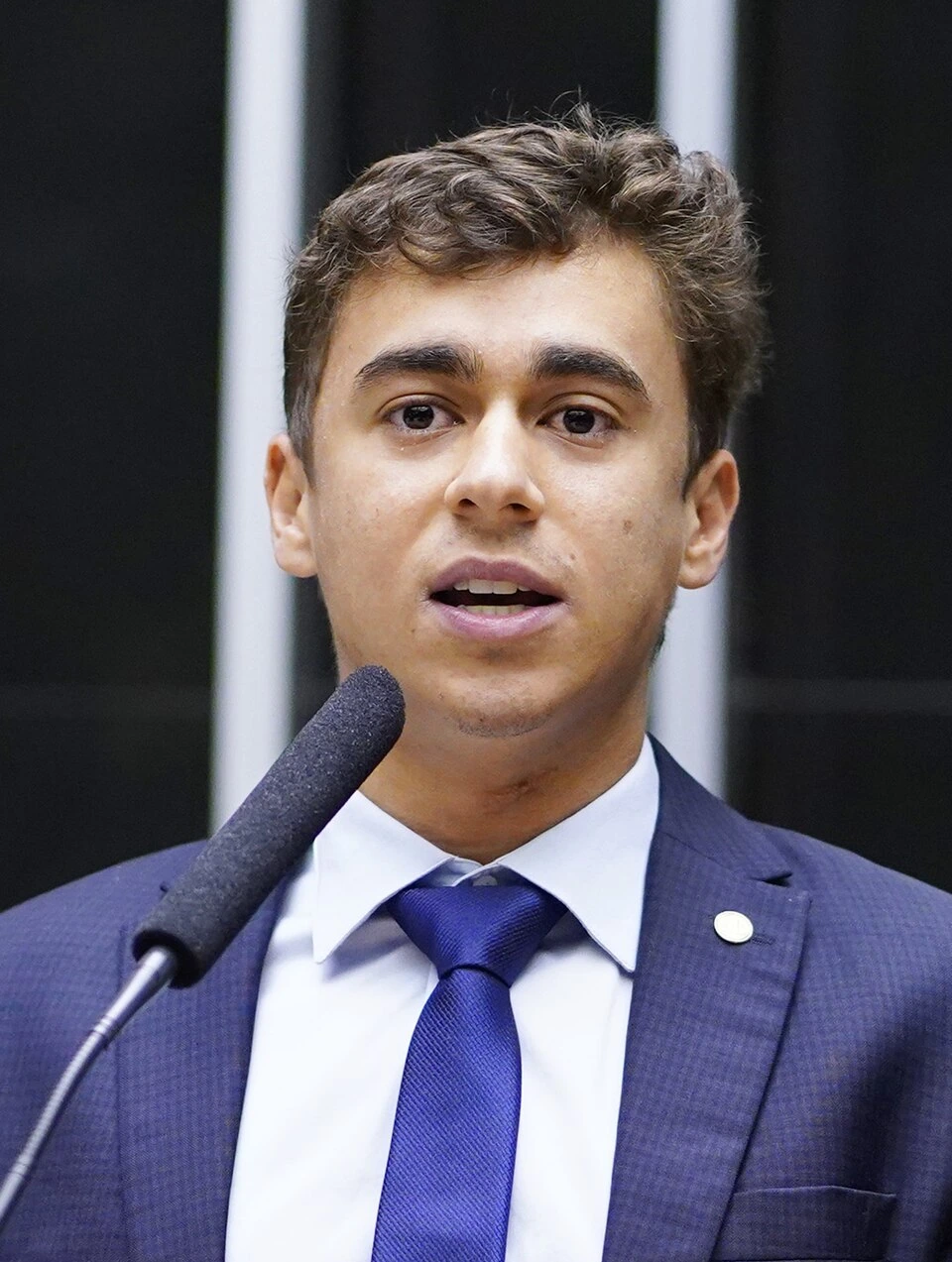 Nikolas Ferreira de Oliveira speaking at a podium in a suit and tie
