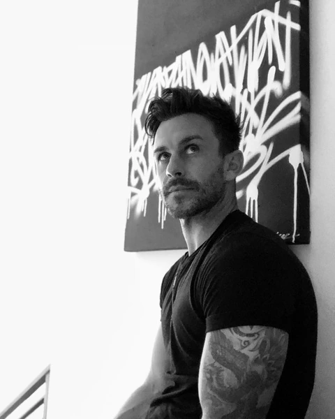 Black-and-white portrait of artist Nicholas Tramontin seated beside his artwork.